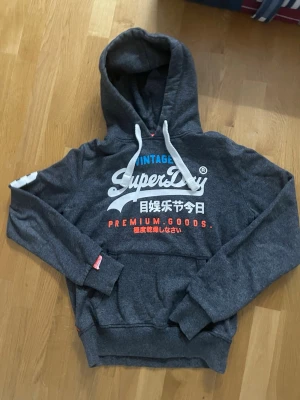 Mörkgrå Superdry hoodie med tryck - Mörkgrå hoodie från Superdry med stor vit och färgglad logga och japansk text på bröstet. Hoodien har vit dragsko i huvan och en klassisk magficka. Tillverkad i mjukt sweatshirtmaterial, perfekt för en avslappnad stil.