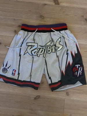 Toronto Raptors basketshorts - Snygga vita basketshorts med Raptors-logga och coolt blixtmönster. Shortsen har snörning i midjan, röda och svarta detaljer samt officiella NBA-märken. Perfekta för dig som gillar streetwear och basket.