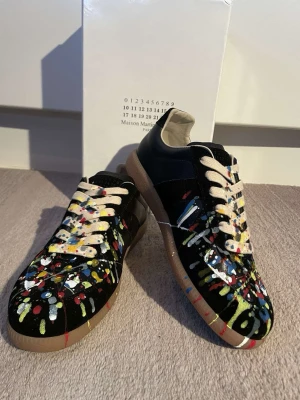 Svarta Maison Margiela skor med färgstänk - Unika svarta Maison Margielas med coola färgstänk i rött, blått, gult, vitt och grönt. Skorna har vita snören och en klassisk rund tå samt platt sula. Perfekta för dig som vill sticka ut med din stil. Jätte bra skick, skriv gärna om ni har frågor. SÄLJER BILLIGT PÅGRUND AV ATT DE FINNS INGEN BOX/TAGS MER OCH LITE ANVÄNDA!!