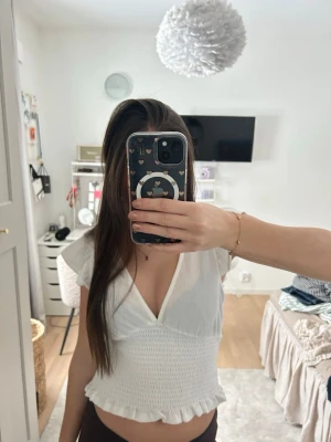 Vit croppad blus med volang - Söt vit blus med v-ringning, korta volangärmar och smockad midja. Toppen är croppad och har en volangkant nedtill. Perfekt för en fräsch och somrig look.