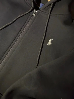 Svart hoodie från Polo Ralph Lauren - Svart hoodie med dragkedja från Polo Ralph Lauren. Klassisk design med vit broderad logga på bröstet, huva med dragsko och två fickor framtill. Perfekt för en chill och stilren look.