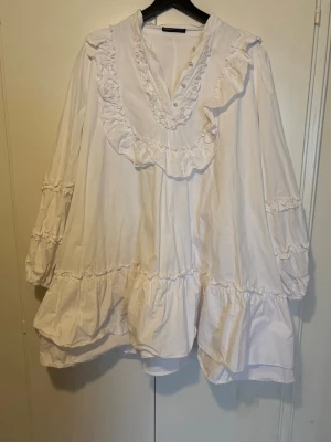 Vit volangklänning med knappar - Söt vit klänning med volanger och dekorativa knappar vid halsringningen. Klänningen har lång ärm med volangdetaljer och en lös, luftig passform. 