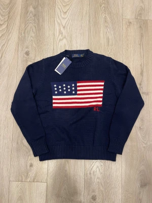 Mörkblå stickad tröja Polo Ralph Lauren - Snygg mörkblå stickad tröja från Polo Ralph Lauren med amerikansk flagga i vitt och rött på bröstet samt RL-broderi. Rund hals och ribbade muddar vid ärmslut och nederkant. Perfekt för dig som gillar klassisk och cool stil.