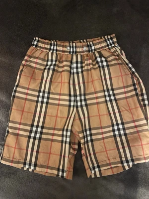 Rutig Burberry shorts - Snygga shorts med klassiskt Burberry rutmönster i beige, svart, vitt och rött. De har elastisk midja med två knappar framtill och fickor på sidorna samt en bakficka. Perfekta för dig som vill sticka ut med en trendig och tidlös look. Snören är borta