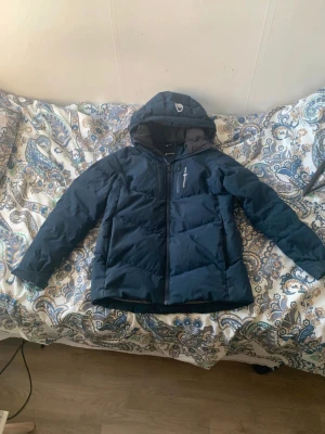 Mörkblå Sail Racing dunjacka - Snygg mörkblå dunjacka från Sail Racing med huva och Sail Antarctica Expedition patch på ärmen STORLEK 170 - 180. Jackan har dragkedja framtill, quiltad design och är långärmad. Perfekt för kalla dagar och har en clean look med diskreta logodetaljer.