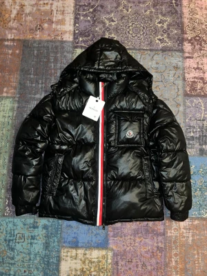 Svart Moncler pufferjacka med huva - Svart pufferjacka från Moncler med glansig finish, huva och ikonisk logga på bröstfickan. Jackan har en rödvit detalj längs dragkedjan, praktiska sidofickor och elastiska muddar. Perfekt för kalla dagar när du vill ha både stil och värme. Jackan är också helt ny och har NFC 