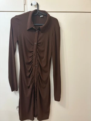 Brun skjortklänning med rynk framtill - Snygg brun skjortklänning med långa ärmar och knappar hela vägen framtill. Klänningen har en cool rynkdetalj längs knappraden och klassisk krage. Perfekt för dig som gillar stilrena och trendiga plagg med en twist.