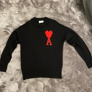 Svart stickad tröja från Ami Paris - Svart stickad tröja från Ami Paris med rund hals och ribbade muddar. På bröstet finns ett stort rött hjärta med bokstaven A. Tröjan har en klassisk passform och är perfekt för dig som gillar stilrena detaljer med en twist.