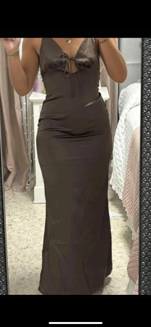 Brun långklänning med snörning - Supersnygg brun långklänning i satinliknande material med smala axelband och snörning både fram och bak. Klänningen har v-ringad urringning och öppen rygg med korsade band, vilket ger en elegant och trendig look. Passformen är slim och klänningen är helt ärmlös.
