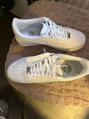 Nike Air Force 1 helvita sneakers - Nike Air Force 1 sneakers i helvit färg med klassisk låg siluett. Skorna har perforerad tå, snörning och platt sula med AIR-detalj på sidan. Tillverkade i skinn med vita skosnören och diskret Nike-logga på sidan. Perfekta för dig som gillar clean och stilren look.