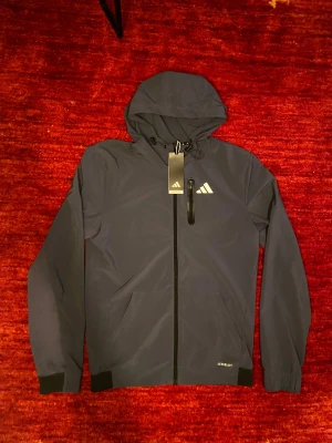 Mörkblå vindjacka från Adidas - Helt ny jacka, aldrig använd och med originalprislapp kvar. Modern och stilren modell som passar perfekt för både vardag och träning. Jackan är bekväm, lätt och praktisk med huva och dragkedja framtill. I absolut nyskick.