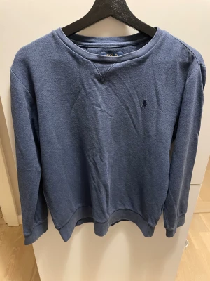Blå sweatshirt från Polo Ralph Lauren - Snygg blå sweatshirt från Polo Ralph Lauren med klassisk rund hals och diskret broderad logga på bröstet. Tröjan har långa ärmar och ribbade muddar vid ärmslut och nederkant. Perfekt för chill dagar och enkel att matcha med jeans.