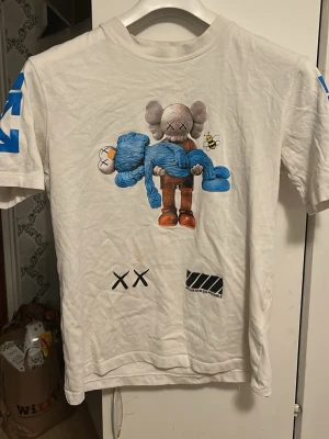 Vit KAWS x Off-White t-shirt - Säljer en vit t-shirt från Off-White med KAWS-motiv fram och bak. Trycket visar KAWS-figurer i blått, grått och gult, samt Off-White-logga och XX-detaljer. T-shirten har korta ärmar med blå tryck på båda ärmarna. Materialet är mjuk bomull.