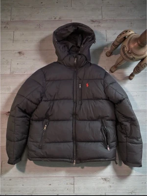 Svart pufferjacka från Polo Ralph Lauren - Svart pufferjacka från Polo Ralph Lauren med röd broderad logga på bröstet. Jackan har huva, tre dragkedjeförsedda fickor och en klassisk quiltad design. Perfekt för kalla dagar och riktigt snygg streetstil.