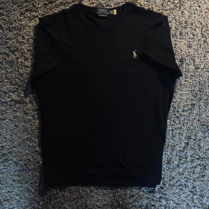 Svart t-shirt från Polo Ralph Lauren - Svart t-shirt från Polo Ralph Lauren med klassisk rund hals och liten broderad logga på bröstet. Modellen är custom slim fit och tillverkad i 100% bomull för skön känsla. Perfekt för dig som gillar stilrena och tidlösa plagg.