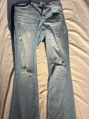 Ljusblå bootcut jeans med slitningar - Säljer ett par ljusblå jeans med bootcut-modell och snygga slitningar på benen. Jeansen har klassisk femficksdesign och är tillverkade i denim. Perfekta för dig som gillar en avslappnad och trendig look.