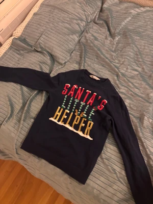 Mörkblå jultröja Santa's Little Helper - Rolig mörkblå jultröja med texten 'Santa's Little Helper' i rött, grönt och guld. Tröjan har långa ärmar och ribbade muddar. Perfekt för dig som vill sticka ut på julfirandet med en färgglad och festlig look.