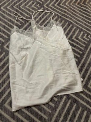 Vit linnetopp med spetsdetaljer - Säljer en vit linnetopp med tunna axelband och vacker spets längs halsringningen. Toppen har en mjuk, slät yta och är lätt och luftig, perfekt för varma dagar. Spetsen ger en extra touch av stil och gör toppen superfin att styla med jeans eller kjol.