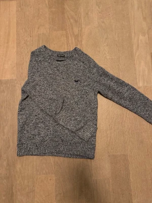 Gråmelerad stickad tröja Hollister XS - Säljer en gråmelerad stickad tröja från Hollister i ullblandning. Tröjan har rund halsringning, lång ärm och en liten broderad logga på bröstet. Perfekt för kyliga dagar och enkel att matcha med jeans eller kjol.