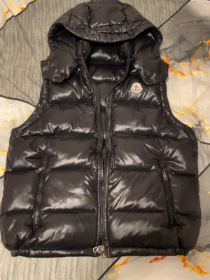 Svart dunväst från Moncler med huva - Svart, glansig dunväst från Moncler med avtagbar huva och två fickor med dragkedja. Västen har ett quiltat mönster och klassisk Moncler-logga på bröstet. Insidan har en cool serietidningsdetalj. Perfekt för dig som vill ha en snygg och varm look.