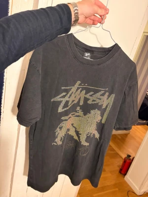 Svart Stüssy Tokyo t-shirt  - Svart lätt urtvättad t-shirt från Stüssy med coolt lejonmotiv och logga i prickmönster på framsidan. Nästan aldrig använd och köpt i Japan för några år sedan. Storlek L men passar M också. 
