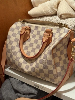 Louis Vuitton Damier handväska beige - Superfin handväska i klassiskt Damier-mönster. Väskan är beige med ljusbruna detaljer och guldiga metalldelar. Den har både handtag och axelrem samt dragkedja upptill. Materialet är canvas med skinn och insidan är ljus. Liten smink fläck i insidan. INTE ÄKTA 