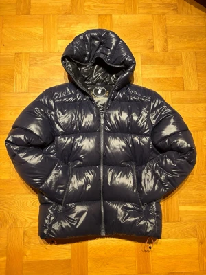 Mörkblå pufferjacka från Save The Duck - Säljer en mörkblå pufferjacka från Save The Duck med glansig finish och huva. Jackan är helt animal free och tillverkad i syntetmaterial. Den har dragkedja framtill som går hela vägen, två sidofickor och coola detaljer på insidan med tryck. Perfekt för dig som vill ha en schysst och varm jacka. Om du undrar något hör bara av dig 