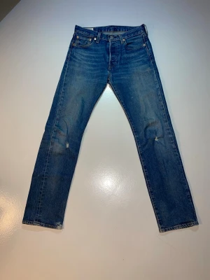 Levi's 501 blå jeans W28 L32 - Klassiska Levi's 501 jeans i blå tvätt med snygga, fabriksgjorda slitna detaljer och raka ben. Jeansen har fem fickor, knappgylf och det ikoniska läderpatchen bak i midjan. Perfekt passform och tidlös stil, tillverkade i robust bomullsdenim. Kan tvättas innan skickas.