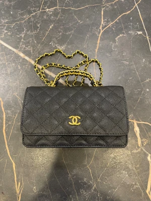 Svart quiltad Chanel axelväska - Supersnygg svart axelväska från Chanel med klassiskt quiltat mönster och guldfärgad kedja. Väskan har det ikoniska CC-emblemet i guld på framsidan och praktiskt fack med dragkedja inuti. Perfekt storlek för det viktigaste och riktigt lyxig vibe.