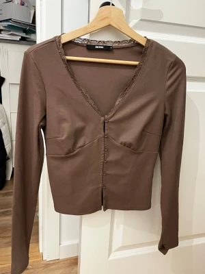 Brun kofta med spets från Bik Bok - Supersnygg brun kofta från Bik Bok i storlek S. Den har v-ringad hals med spetsdetaljer, knappar framtill och långärmad passform. Materialet är mjukt och stretchigt, perfekt för lager på lager. Sitter normal och har en feminin look med fina sömmar över bysten. (Den sista knappen går ej att knäppa)