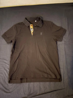 Burberry piké - Säljer en svart pikétröja från Burberry med klassisk rutig knappslå i beige, svart och rött. Tröjan har korta ärmar, krage och diskret broderad B-logga på bröstet. Tillverkad i bomull för en skön och stilren look.