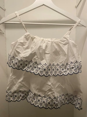 Vit broderad linnetopp från LMTD - Supersöt vit linnetopp från LMTD i storlek XS med tunna axelband och broderade blå blommor längs volangkanterna. Toppen har dubbla lager med vågig kant. Materialet är mjuk bomull och ger en luftig känsla.