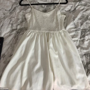 Vit spetsklänning med smala axelband - Supersöt vit kortklänning med smala axelband och spetsdetaljer upptill. Kjolen är lätt utställd och ger en flowy känsla. Klänningen har dragkedja i ryggen och ett romantiskt spetsmönster på överdelen. Perfekt för skolavslutning eller studenten!