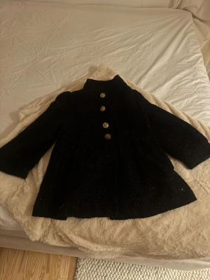 Svart rock med guldknappar - Snygg svart rock med rak passform och höga axlar. Jackan har fyra stora guldknappar framtill och en lite glittrig yta. Materialet känns tjockt och lite grovt, perfekt för kyliga dagar. Ståkrage och långa ärmar ger en stilren vibe.