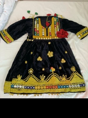 Svart traditionell klänning med broderi - Unik svart klänning med färgglada broderier och spegeldekorationer i gult, rött, blått och grönt. Klänningen har trekvartsärm, v-ringning och detaljer som blommor, paljetter och en stor röd tygblomma på bröstet. Perfekt för dig som vill sticka ut med traditionell stil. Storlek 5-6 år. Arabisk klänning 