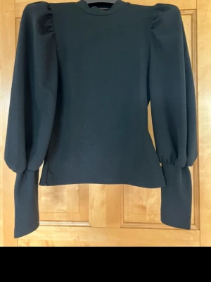 Svart blus med puffärm från SHEIN - Svart blus från SHEIN i storlek S med höga puffärmar och rund hals. Materialet är strukturerat och känns lite tjockare, vilket ger en cool siluett. Ärmarna är långa och har extra volym vid axlarna för en trendig look.