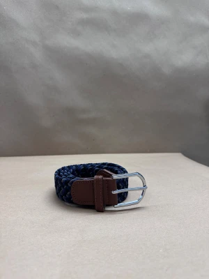 Blå flätad bälte med bruna detaljer - Snyggt flätat bälte i marinblått och mörkblått med bruna skinn-detaljer vid spännet. Bältet har ett klassiskt silverfärgat metallspänne och är gjort i ett mjukt, flexibelt material som ger en cool och modern look. Perfekt för att lyfta din outfit.
