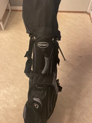 Svart golfbag Open med klubbor - Hej säljer ett golf sett med 9klubber dom är andvända. Golf väskan får man med när man köper hela sättet. Annars ser bar ut fick det av min kommpis men har ingen användning för dem.