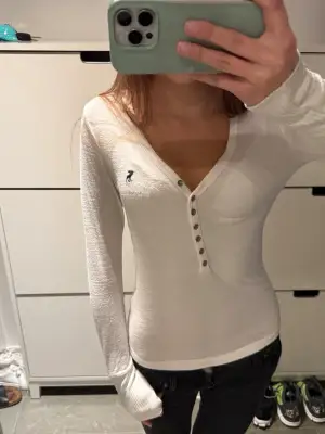 Henley. Snygg vit långärmad topp från Abercrombie & Fitch med åtta knappar framtill. Tröjan har ribbade muddar och ett broderat mörkblått älglogo på bröstet. Mjuk och tunn bomullskvalitet som är perfekt för lager-på-lager.