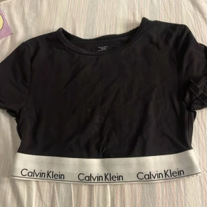 Svart croppad topp Calvin Klein S - Svart croppad topp från Calvin Klein med korta ärmar och rund hals. Nedtill finns en bred vit resår med Calvin Klein-logga i svart. Toppens material är mjukt och stretchigt, perfekt för en chill och sportig look.
