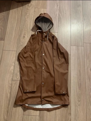 Brun regnjacka med huva - Snygg brun regnjacka med huva och vita snören. Jackan har knappar framtill och är längre baktill för extra skydd mot regn. Perfekt för blöta dagar och har en stilren look med enkel design.