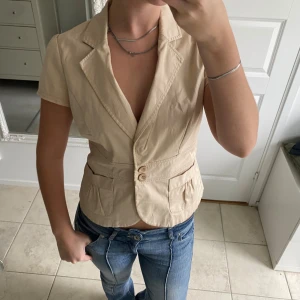 Beige kortärmad kavaj med knappar - Snygg beige kavaj med korta ärmar och två knappar framtill. Jackan har två fickor och markerad midja för en cool siluett. Perfekt att styla med jeans eller kjol för en trendig look.