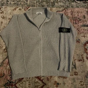 Grå stickad kofta från Stone Island - Säljer en grå stickad kofta från Stone Island med dragkedja framtill och ribbad struktur. Klassisk svart patch med logga på vänster ärm. Perfekt för dig som gillar clean och stilren streetwear. Koftan har lång ärm och hög krage.