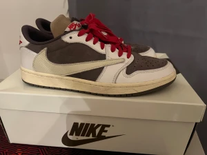 Nike Air Jordan 1 Low OG Travis Scott - Nike Air Jordan 1 Low OG Travis Scott med omvänd vit swoosh, bruna och vita paneler i skinn och mocka samt röda snören. Ikonisk sneaker med rund tå och platt sula, detaljerad med Air Jordan-logga på hälen och Travis Scott branding. Perfekt för dig som vill sticka ut.