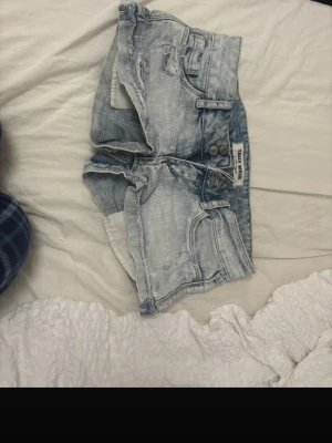 Ljusblå lågmidjade jeans shorts - Säljer ett par ljusblå denimshorts  med slitna detaljer och klassisk femficksdesign. Shortsen har låg midja, två knappar framtill och råa kanter för en cool vibe. Perfekta för varma dagar och chill häng.