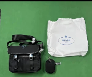 Svart axelväska från Prd - Snygg svart axelväska från prd med klassisk trekantig logga framtill. Väskan har justerbar axelrem, två framfickor med spännen och dragkedja, samt medföljande liten accessoar. Materialet är slitstark nylon med detaljer i skinn och metall. Kan skicka 7-18 mer bilder privat 