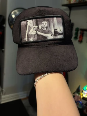 Svart velvet keps med SampeV2 Langa bican  - Svart truckerkeps med mesh baktill och justerbar snapback. Framtill finns ett svartvitt tryck med motiv av en person som flexar armen. Kepsen har en klassisk böjd skärm och är tillverkad i syntetmaterial.