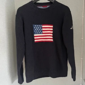 Mörkblå stickad tröja från Gant - Snygg mörkblå stickad tröja från Gant med amerikansk flagga framtill och broderad text 'GANT NEW HAVEN 1949' på ärmen. Tröjan har rund halsringning och långa ärmar, perfekt för en preppy look.