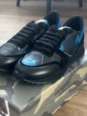 Valentino Garavani Rockrunner sneakers - Säljer ett par Valentino Garavani Rockrunner sneakers i svart och blått med coolt kamouflagemönster. Skorna har snörning, rund tå och detaljer i både mocka och skinn. Sulan är räfflad för extra grepp och boxen har matchande camo-print. Perfekta för dig som vill sticka ut med lyxig streetstyle.
