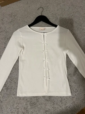 Vit långärmad topp med rosetter från Nelly - Supersöt vit ribbad topp från Nelly i storlek XS. Långärmad med rund hals och snygga öppna detaljer framtill som hålls ihop av flera små rosetter. Materialet är mjukt och stretchigt, perfekt för dig som gillar en feminin och trendig look.